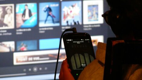 Photo d'une personne écoutant de la musique avec son smartphone avec en fond des jaquettes d'albums sur grand écran